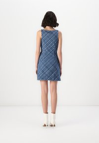 Majica brez rokavov iz denima z modrim karirastim vzorcem, prilegajoča se zasnova in ravni rob, v kombinaciji z belimi gležnjarji. Prikazan zadnji pogled.