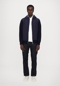 Navy zip-up hoodie med ribbad mudd, bärs över en vit polo, ihop med mörka denimjeans och vita sneakers, stående mot en enfärgad bakgrund.