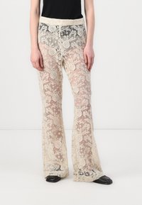 Pantalons évasés en dentelle crème avec des motifs floraux intriqués, dotés d'une large ceinture élastique et d'une apparence transparente et texturée.