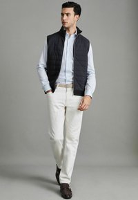 Jachetă de quiltată neagră peste o cămașă butonată bleu deschis, combinată cu pantaloni albi și pantofi din piele maro cu două catarame.
