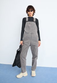 Gray denim overalls with adjustable straps and front pocket, paired with a long-sleeve black top and beige sneakers, holding a black tote bag.