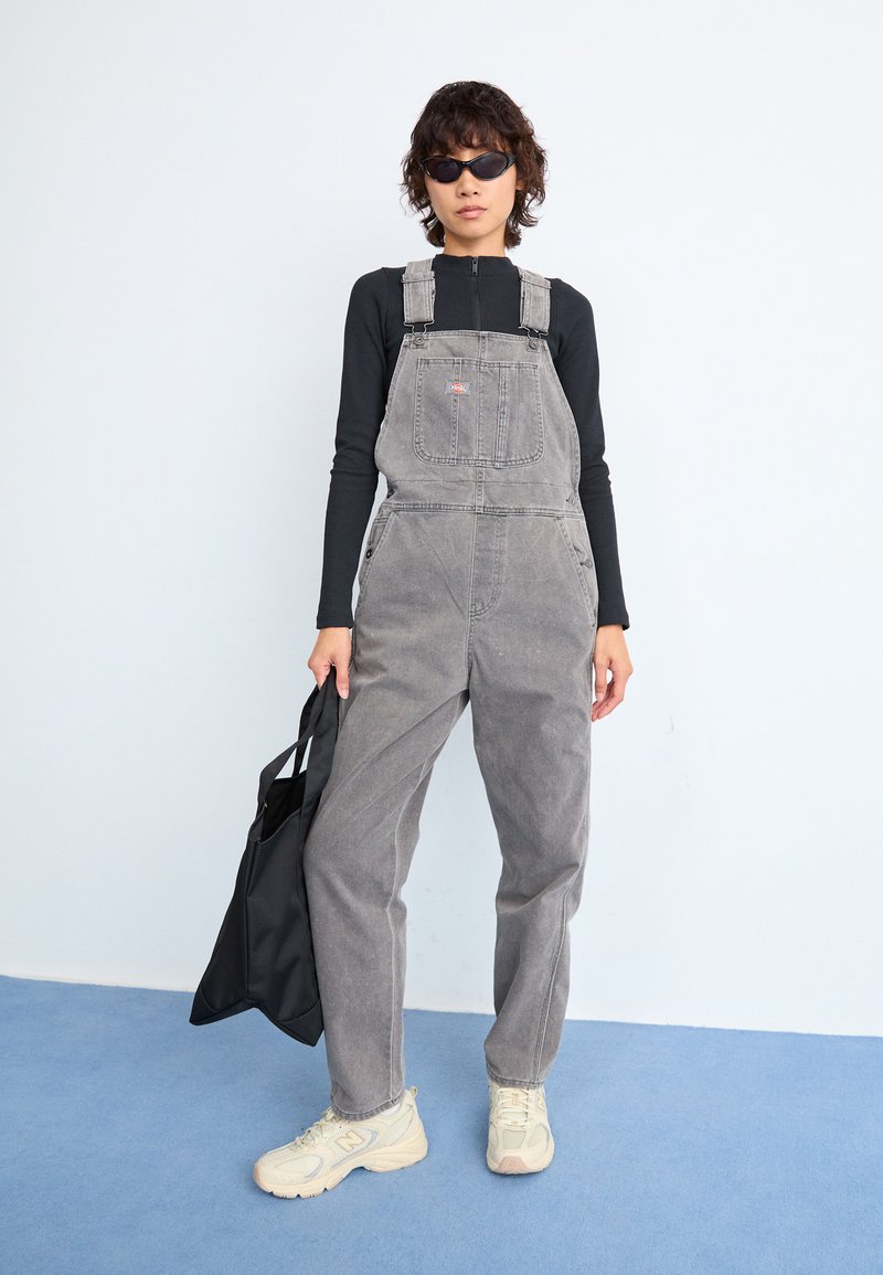 Graue Denim-Latzhose mit verstellbaren Trägern und Fronttasche, kombiniert mit einem langärmeligen schwarzen Oberteil und beigen Sneakers, während sie eine schwarze Tragetasche hält.