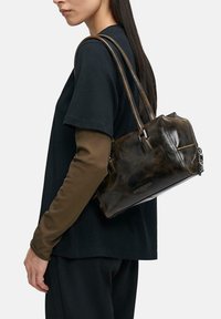 Frau mit schwarzem Shirt und braunen, langen Ärmeln trägt eine glänzende, dunkelbraune Lederschultertasche mit doppelten Riemen.