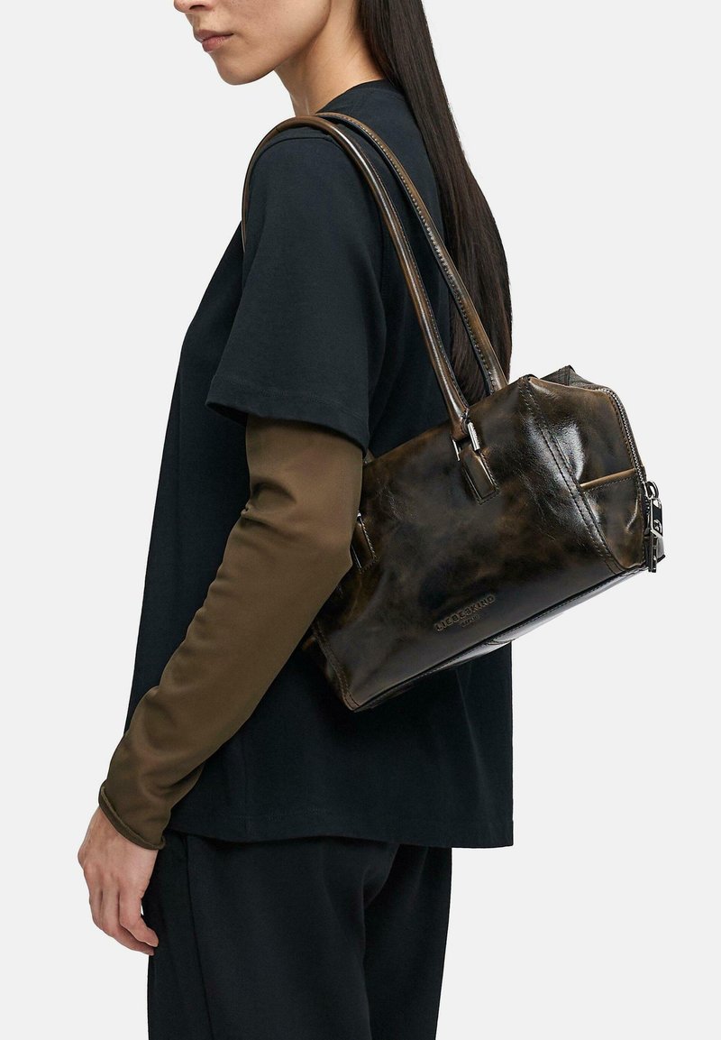 Frau mit schwarzem Shirt und braunen, langen Ärmeln trägt eine glänzende, dunkelbraune Lederschultertasche mit doppelten Riemen.