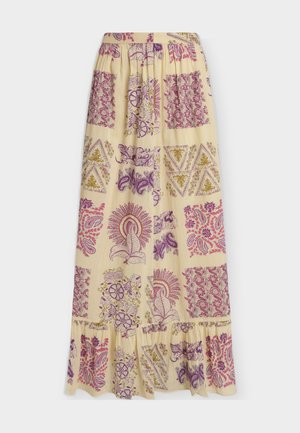 Femme debout portant une robe maxi jaune sans manches avec des motifs floraux violets et roses et des sandales noires sur fond blanc.