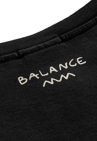 Czarna bawełniana tkanina z żebrowanym dekoltem. Na plecach widnieje napis "BALANCE" w kremowym, falistym stylu. Teksturowana powierzchnia.