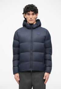Námornícka prešívaná bunda s prešitými baffle vzormi, plný zips, kapucňa so sťahovacou šnúrkou a malým logom na hrudi. Ľahká a izolačná.