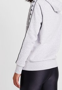 Hoodie grigio con cappuccio e dettagli neri, caratterizzato da un design con logo lungo le maniche. Tessuto morbido con polsini e orlo a costine.