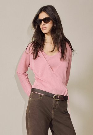 Kvinde med langt mørkt hår iført mørke solbriller, en lyserød omslagstrøje over en lyserød top, mørkebrune bukser og et sort bælte med en metalspænde.