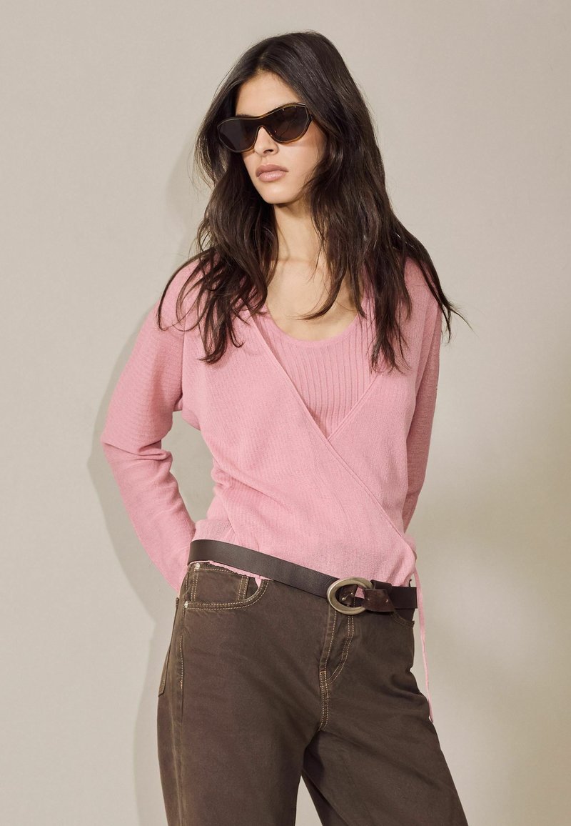 Žena s dlhými tmavými vlasmi má na sebe tmavé slnečné okuliare, ružový zavinovací sveter cez ružový top, tmavohnedé nohavice a čierny opasok s kovovou sponou.