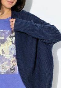 Marinblå stickad kofta med texturerade ärmar, bärs över en blå t-shirt med grafiskt tryck som har mystiska inslag och text.