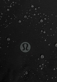 Čierna tkanina s rozptýlenými kvapkami vody, so zvýrazneným šedým logom v strede. Textúra vyzerá hladko s jemným leskom.