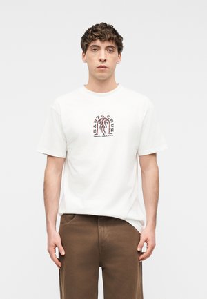 Tânăr în picioare, în față, purtând un tricou alb Santa Cruz Skateboards și pantaloni maro, pe un fundal simplu.