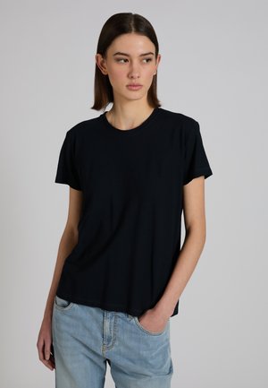 Vrouw met steil bruin haar, gekleed in een effen zwart T-shirt en lichtblauwe spijkerbroek, staand met één hand in de zak, kijkt zijwaarts.