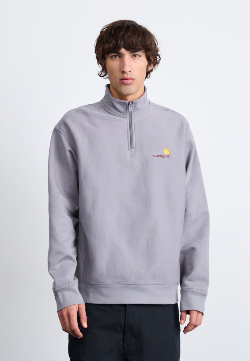 Pull en quarter-zip de couleur gris clair, fabriqué dans un tissu doux avec des poignets et un ourlet côtelés. Présente un petit logo brodé sur la poitrine.