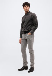 Νεαρός άνδρας στέκεται με το χέρι στην τσέπη, φοράει σκούρο γκρι πουκάμισο με κουμπιά, ανοιχτό γκρι τζιν, μαύρη ζώνη και μαύρα δερμάτινα μποτάκια αστραγάλου.