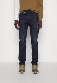 Vaqueros de mezclilla de un azul oscuro con corte recto. Combinados con una camisa de manga larga marrón y zapatillas verdes con acentos en beige.
