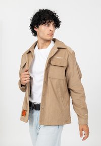 Jasnobrązowa kurtka z wykroju w kordówkę z zapięciem na guziki, dwoma przednimi kieszeniami oraz kieszenią na suwak na rękawie. Nosi ją na białym T-shircie.