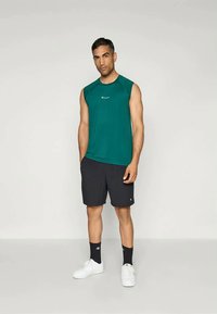 Mörkgrön ärmlös sporttröja med logotyp, tillsammans med svarta shorts. Bärs med vita sneakers och svarta ankelsockor.