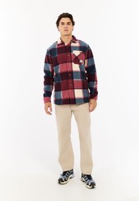Man staat in beige broek, zwart-grijze sneakers, en een rood, blauw en crèmekleurig geruit overhemd met een borstzakje.