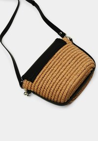 Tkaná béžová kabelka s černým syntetickým koženým klopou a popruhem, která má obdélníkový tvar a texturovaný povrch. Kovové akcenty ve stříbrné barvě.