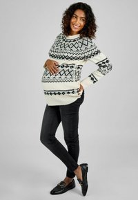 Strickmaternitätspullover in Beige mit schwarzen geometrischen Mustern, kombiniert mit schwarzen Skinny Jeans und schwarzen Slippern. Glatte Textur.