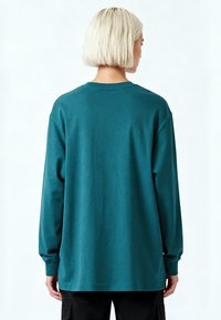 Personne aux cheveux blonds courts portant un chemisier manches longues bleu sarcelle et un pantalon noir, vue de dos sur un fond blanc uni.