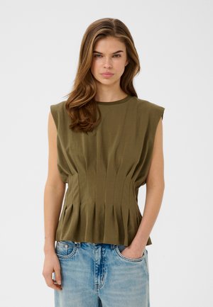Ung kvinde med langt brunt hår iført en ærmeløs olivengrøn plisseret top og lyseblå jeans, stående med den ene hånd i lommen.