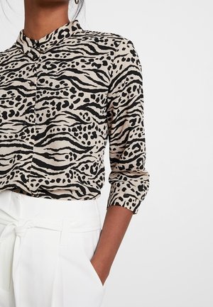 Vrouw draagt een blouse met lange mouwen, zwart-beige dierenprint en een wit, hooggetailleerd broek met een geknoopt lint.