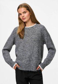 Femme aux longs cheveux châtains clairs portant un pull en tricot gris et un pantalon noir, se tenant avec les mains dans les poches devant un fond blanc.