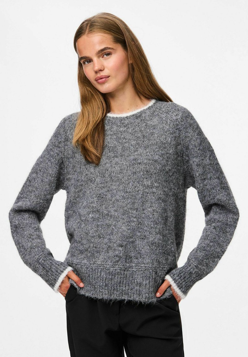 Femme aux longs cheveux châtains clairs portant un pull en tricot gris et un pantalon noir, se tenant avec les mains dans les poches devant un fond blanc.