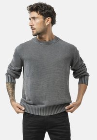Pull en maille gris avec des poignets et un ourlet côtelés, présentant une encolure ronde et une texture douce. Le modèle porte un jean noir ajusté.