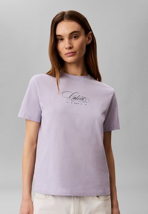 Femme aux cheveux bruns raides portant un t-shirt Calvin violet clair et un pantalon clair sur un fond gris uni.