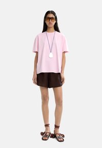 Różowa bawełniana koszulka z krótkim rękawem, brązowe krótkie spodenki oraz brązowe sandały na paski z frędzlami. Dodatki obejmują ogromne bursztynowe okulary przeciwsłoneczne oraz wisiorek.