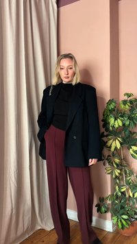 Mujer rubia de pie en el interior, lleva un blazer negro de gran tamaño, top negro y pantalones burdeos, junto a una cortina beige y una planta variegada en maceta.
