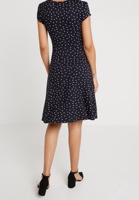 Robe marine à pois blancs, manches courtes, corsage ajusté, jupe évasée et tissu lisse ; portée avec des talons à bride cheville noirs.