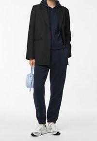 Asymmetrisk svart blazer över en marinblå hoodie, i kombination med marinblå joggers, vita sneakers och en ljusblå handväska. Texturerade tyger.