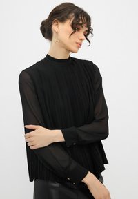 Blusa nera plissettata realizzata in tessuto trasparente con colletto alto, maniche lunghe e dettagli con bottoni dorati ai polsini. Design testurizzato e fluido.