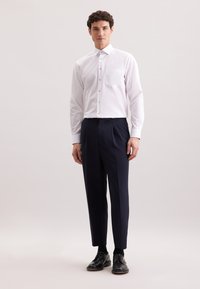 Camicia bianca con bottoni e tasca sul petto, abbinata a pantaloni con pieghe color navy. Il modello indossa scarpe nere, su uno sfondo neutro.