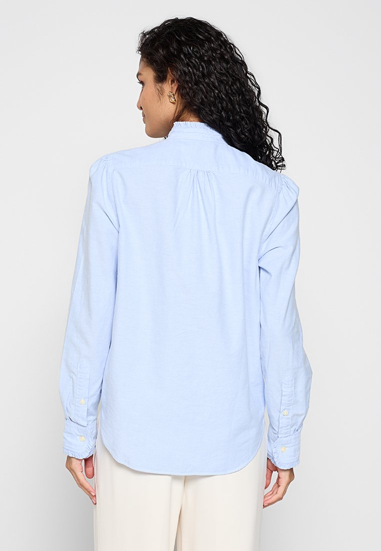 Himmelblaues Langarmshirt aus Baumwolle. Verfügt über einen knöpfbaren Kragen, abgerundetem Saum und Raffung auf der Rückseite. Knöpfe an den Ärmeln.