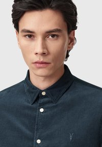 Camicia di velluto a coste blu scuro con colletto button-down, caratterizzata da un logo ricamato discreto e bottoni color crema. Texture liscia.