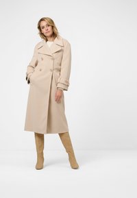 Beige dubbelknäppt kappa med stor krage, knapppdetaljer och uppvikta ärmar. Bärs över en ljus tröja, tillsammans med knähöga beige stövlar.