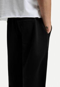 Volné černé kalhoty vyrobené z měkkého materiálu, s bočními kapsami a uvolněným střihem, v kombinaci s bílým topem s krátkým rukávem.