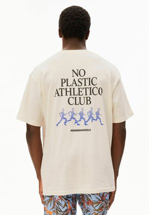Man draagt een beige T-shirt met de tekst "NO PLASTIC ATHLETICO CLUB" en blauwe hardloopfiguren boven het "ARMEDANGELS"-logo, gecombineerd met een korte broek met bloemenprint.
