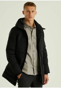 Schwarze Pufferjacke mit Kapuze, Reißverschluss und Seitentaschen. Über einem hellgrauen Hemd mit Knöpfen getragen. Glatte Textur, isoliertes Design.