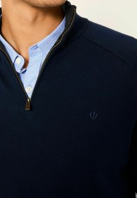Marineblauer Pullover mit Reißverschluss aus weichem Strickmaterial, mit einem dezenten gestickten Logo auf der Brust und einem kontrastierenden Kragen darunter.