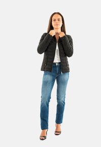 Vrouw die de kraag van een zwart gewatteerd jack aanpast, gekleed in blauwe jeans, een wit shirt en zwarte open teen hakken, staande tegen een effen lichte achtergrond.