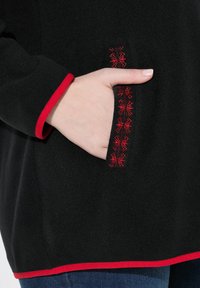 Haut en polaire noir avec des bordures rouges, détails de poche brodés et une poche latérale avec des coutures décoratives.