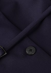 Marineblauw stof met een gladde textuur, voorzien van een zwarte knop met vier gaten. Een stuk stof overlapt en toont een nette naadafwerking.
