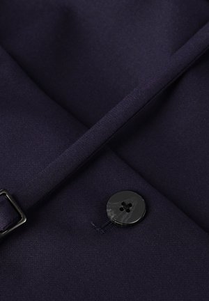 Marineblauw stof met een gladde textuur, voorzien van een zwarte knop met vier gaten. Een stuk stof overlapt en toont een nette naadafwerking.